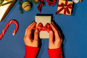 Hands holding a Christmas gift with red ribbon on a blue surface, surrounded by festive decorations.