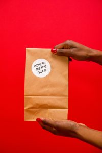 Hands holding a paper bag with "Hope to See You Soon" sticker on a red background.